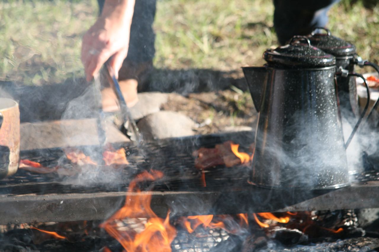 A hand with tongs cooking bacon and coffee over a campfire - Lazy L&B Dude Ranch Wyoming